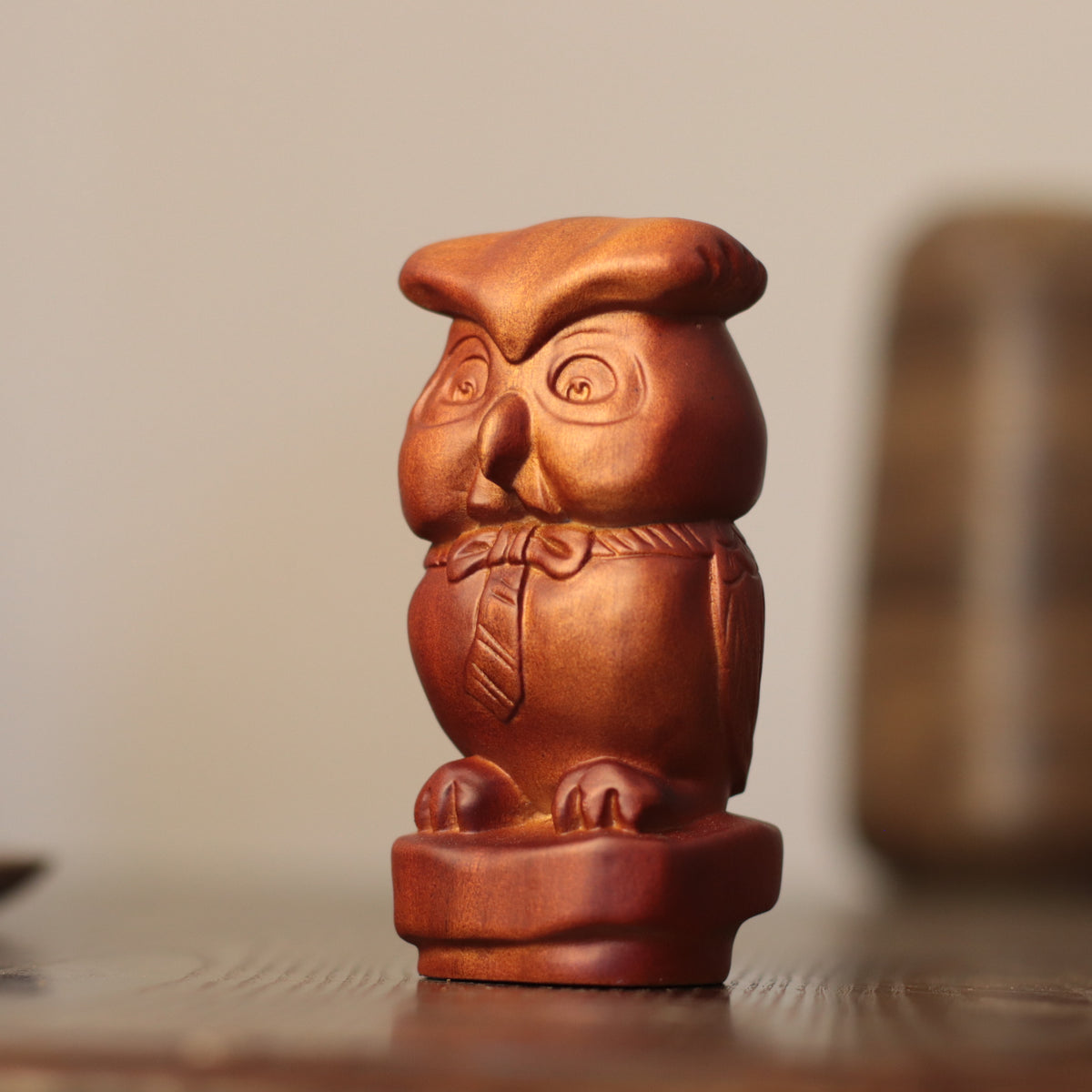 A small carved wooden owl figurine with a warm reddish-brown finish, standing on a matching base against a softly blurred indoor background.
