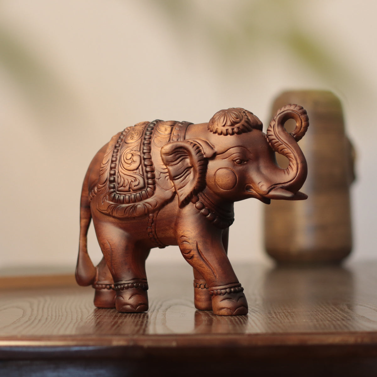 A small carved elephant figurine in warm medium brown wood, standing on a wooden surface with a softly blurred neutral background.