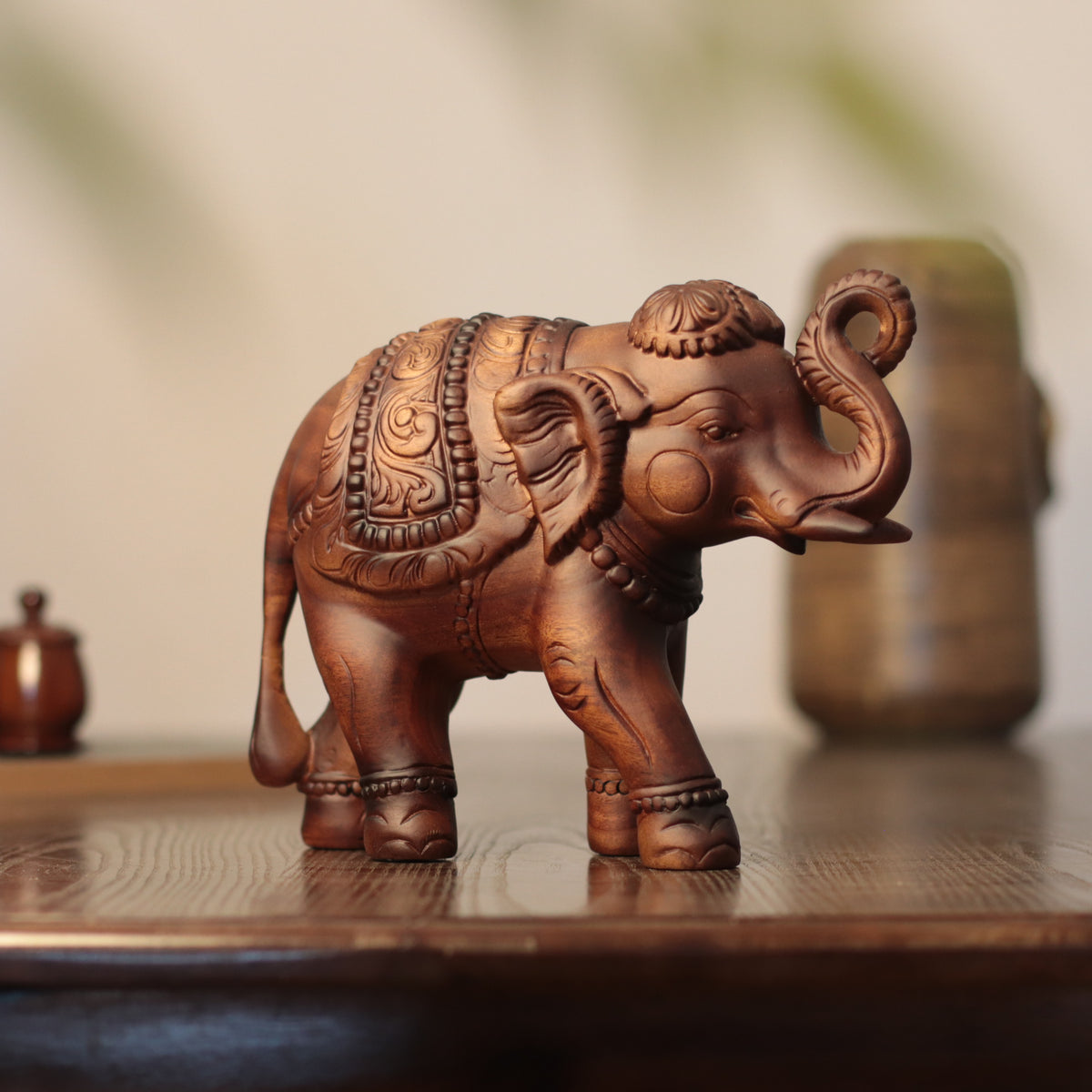 A carved wooden elephant figurine with intricate decorative patterns, displayed on a polished wooden surface with a softly blurred background.