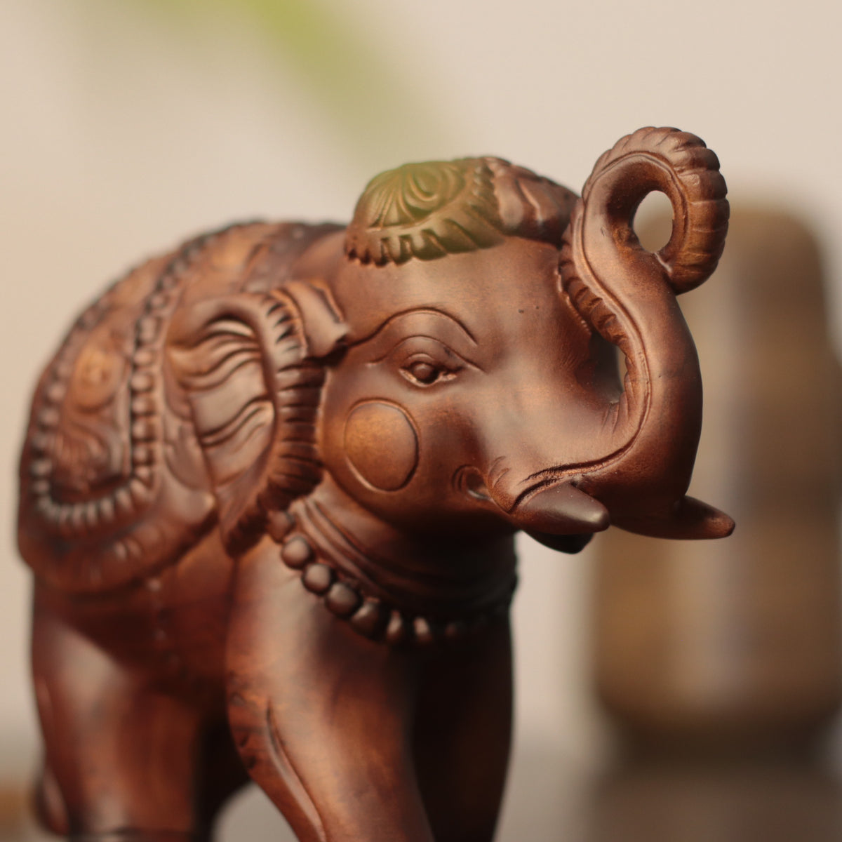 A close-up of a carved elephant figurine with a warm brown finish and subtle green detailing on its head, set against a softly blurred neutral background.