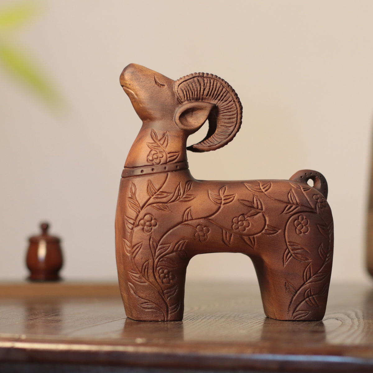 A carved wooden ram figurine with floral engravings, displayed on a wooden surface against a soft beige background.