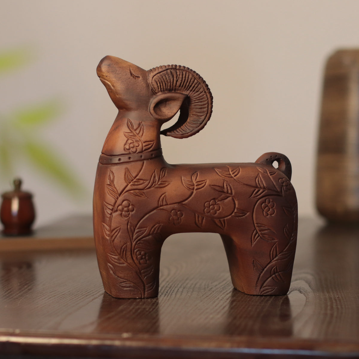 A carved wooden ram figurine with floral engravings, displayed on a dark wooden table against a softly blurred neutral background.