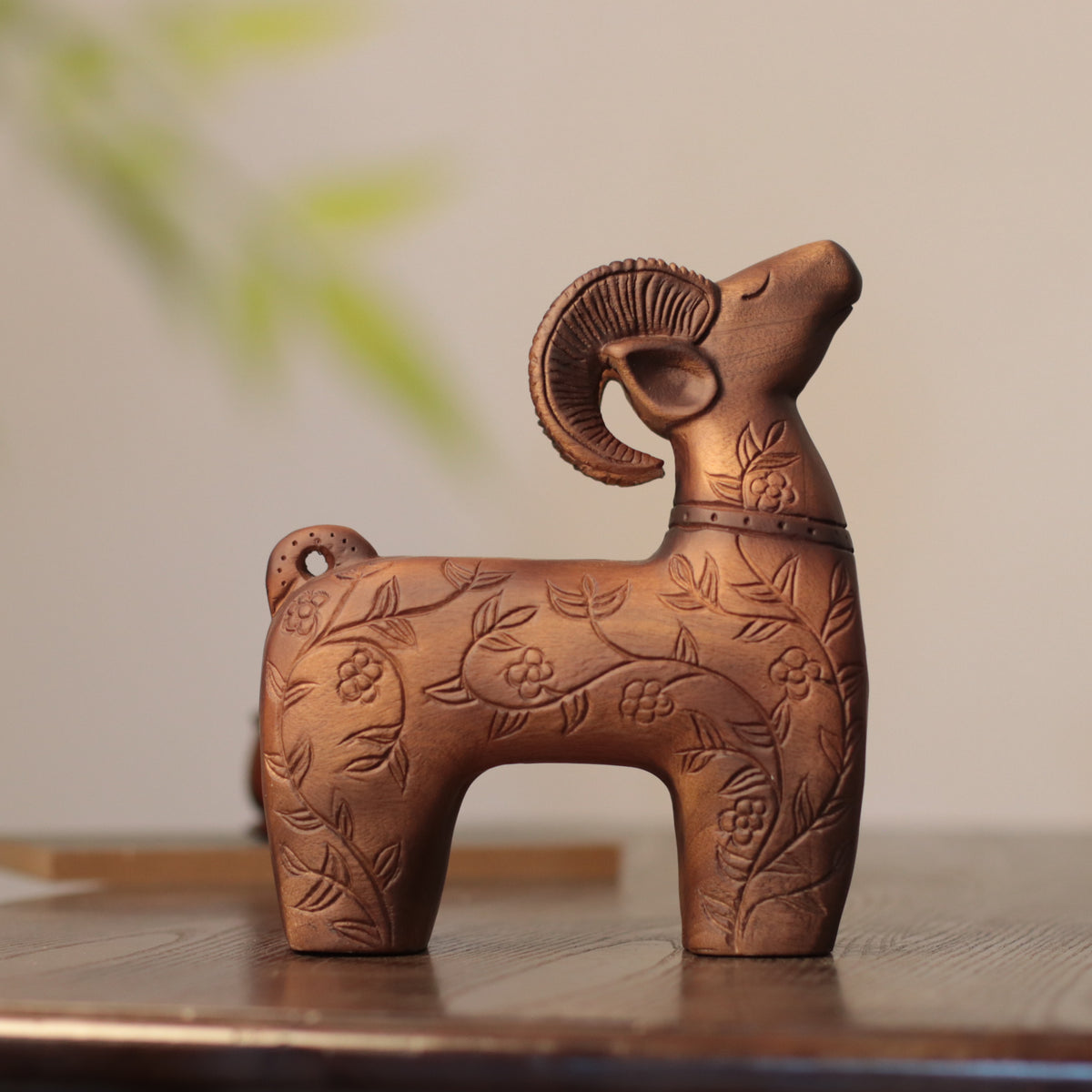 A decorative ram figurine with carved floral patterns, displayed on a wooden surface against a softly blurred beige background.