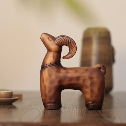 A rustic bronze ram figurine with a curved horn, displayed on a wooden table with a blurred ceramic pot in the background.