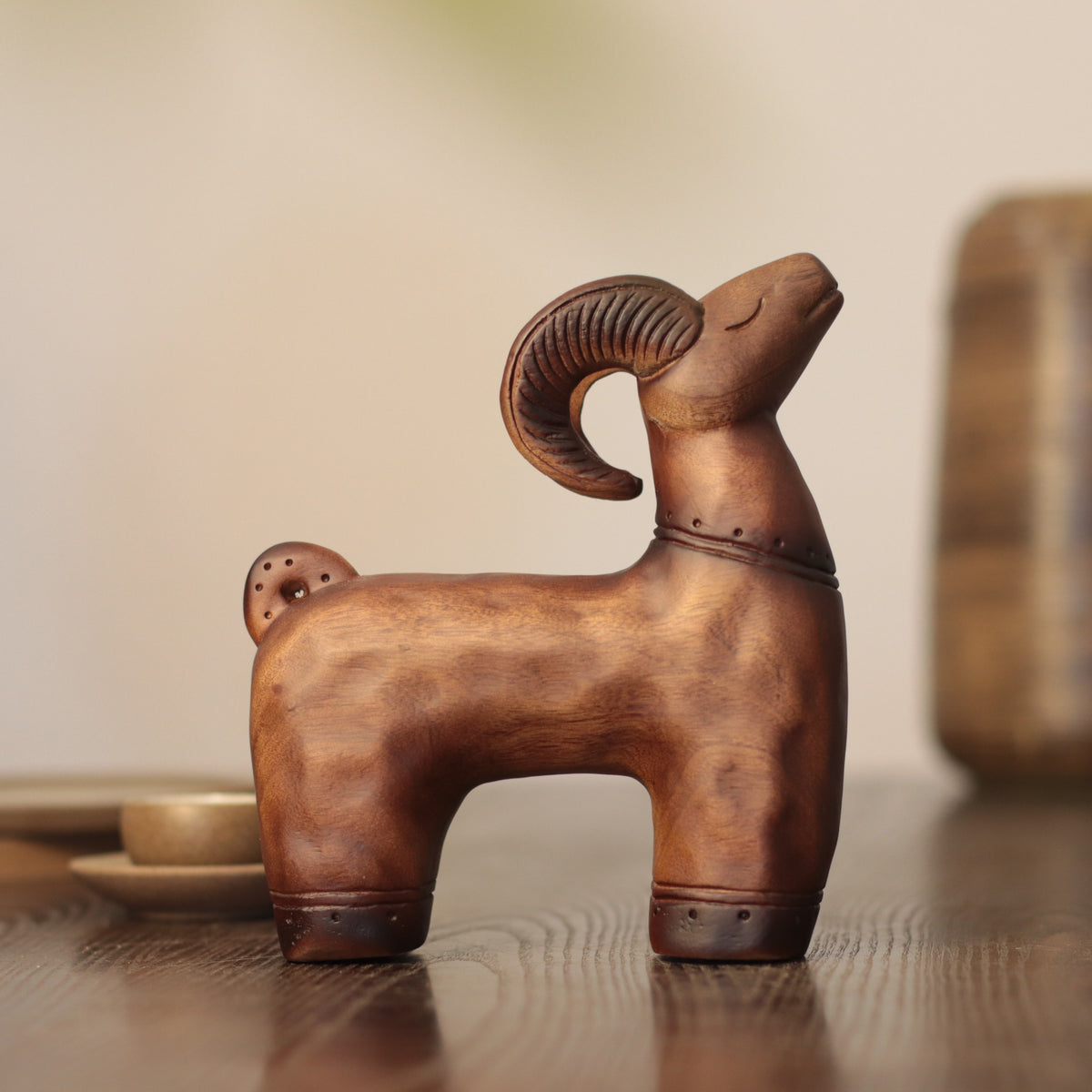 A small carved ram figurine in a warm brown wood or metal finish, standing on a wooden surface with a softly blurred indoor background.
