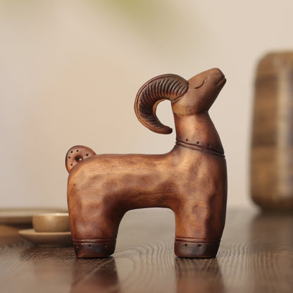 A small carved ram figurine in a warm brown wood or metal finish, standing on a wooden surface with a softly blurred indoor background.