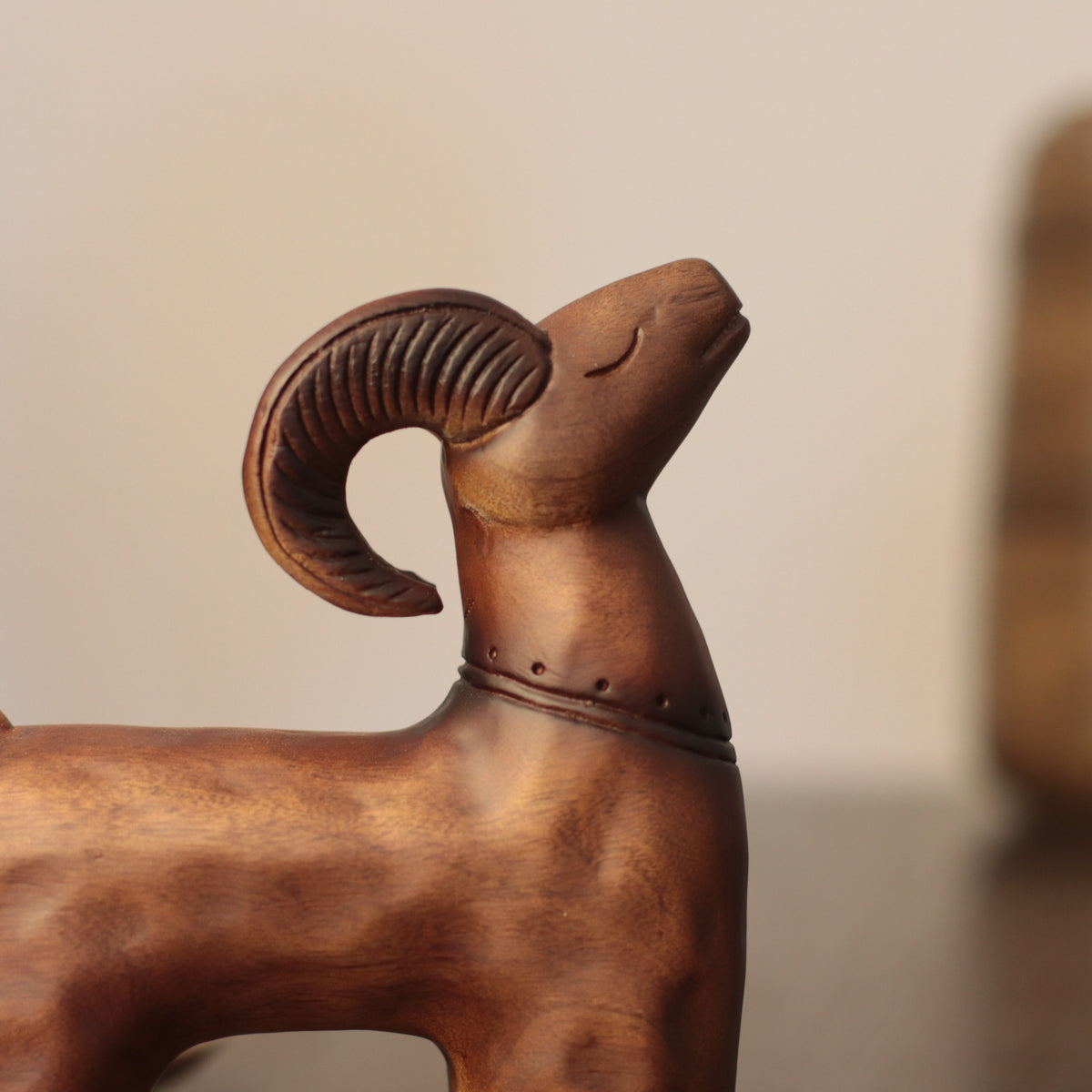 A close-up of a carved wooden ram figurine with curved horns, shown against a softly blurred beige background.