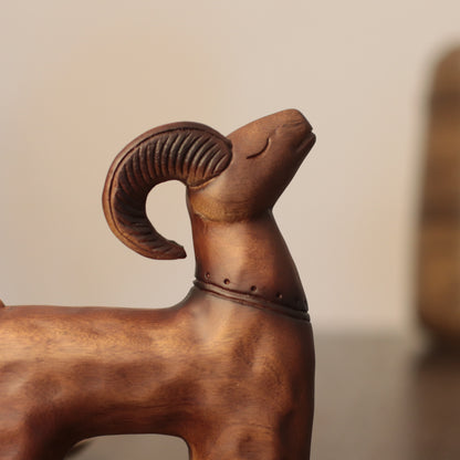 A close-up of a carved wooden ram figurine with curved horns, shown against a softly blurred beige background.