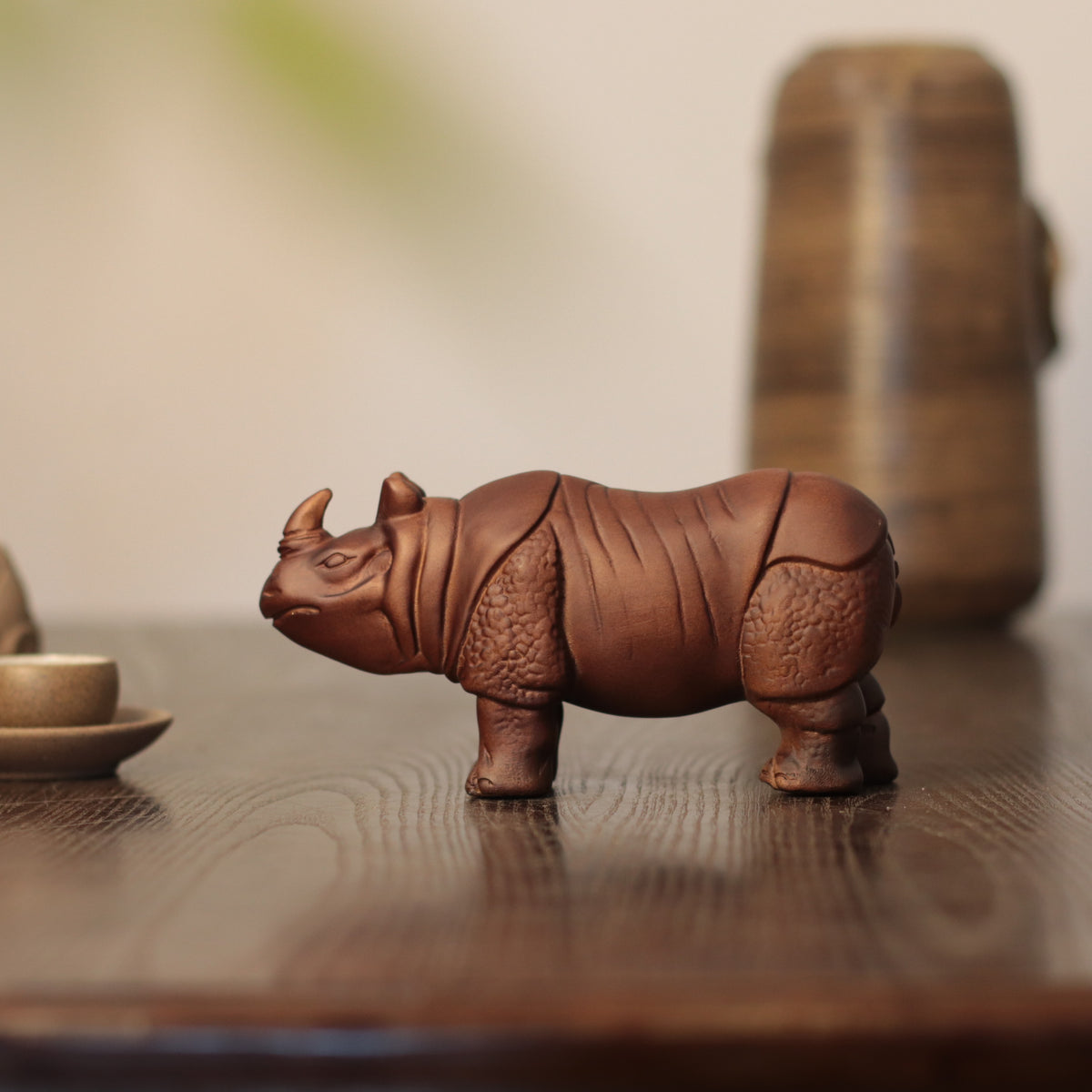 A small brown ceramic rhinoceros figurine displayed on a dark wooden table, with a softly blurred background containing a wooden vessel and a small cup.
