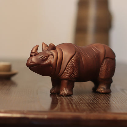 A small brown rhinoceros figurine with detailed textured skin and two horns, standing on a polished wooden surface with a softly blurred background.