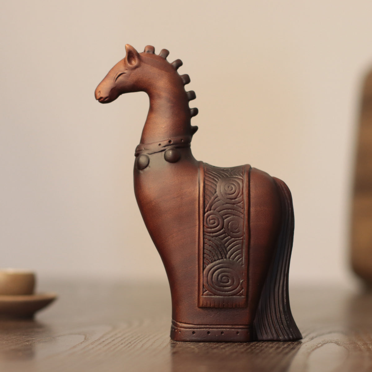 A carved wooden horse figurine with a dark brown finish, featuring an embossed pattern on the side and a decorative saddle, displayed on a wooden surface against a soft beige background.