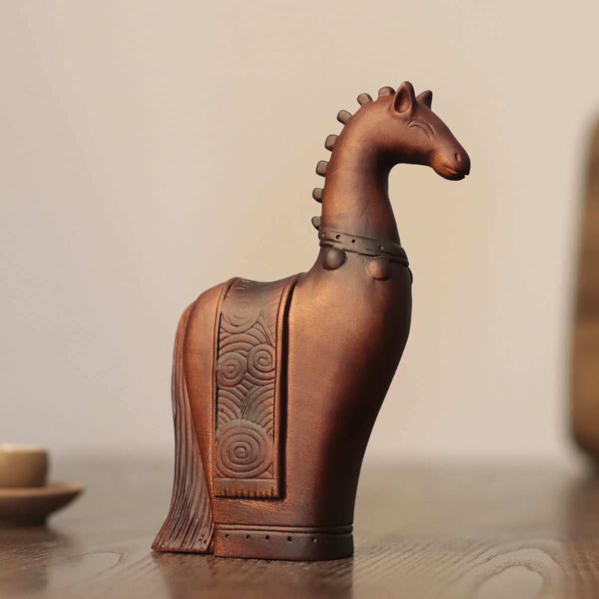 A small sculptural horse figurine in a warm brown tone, standing on a wooden surface against a softly blurred neutral background.