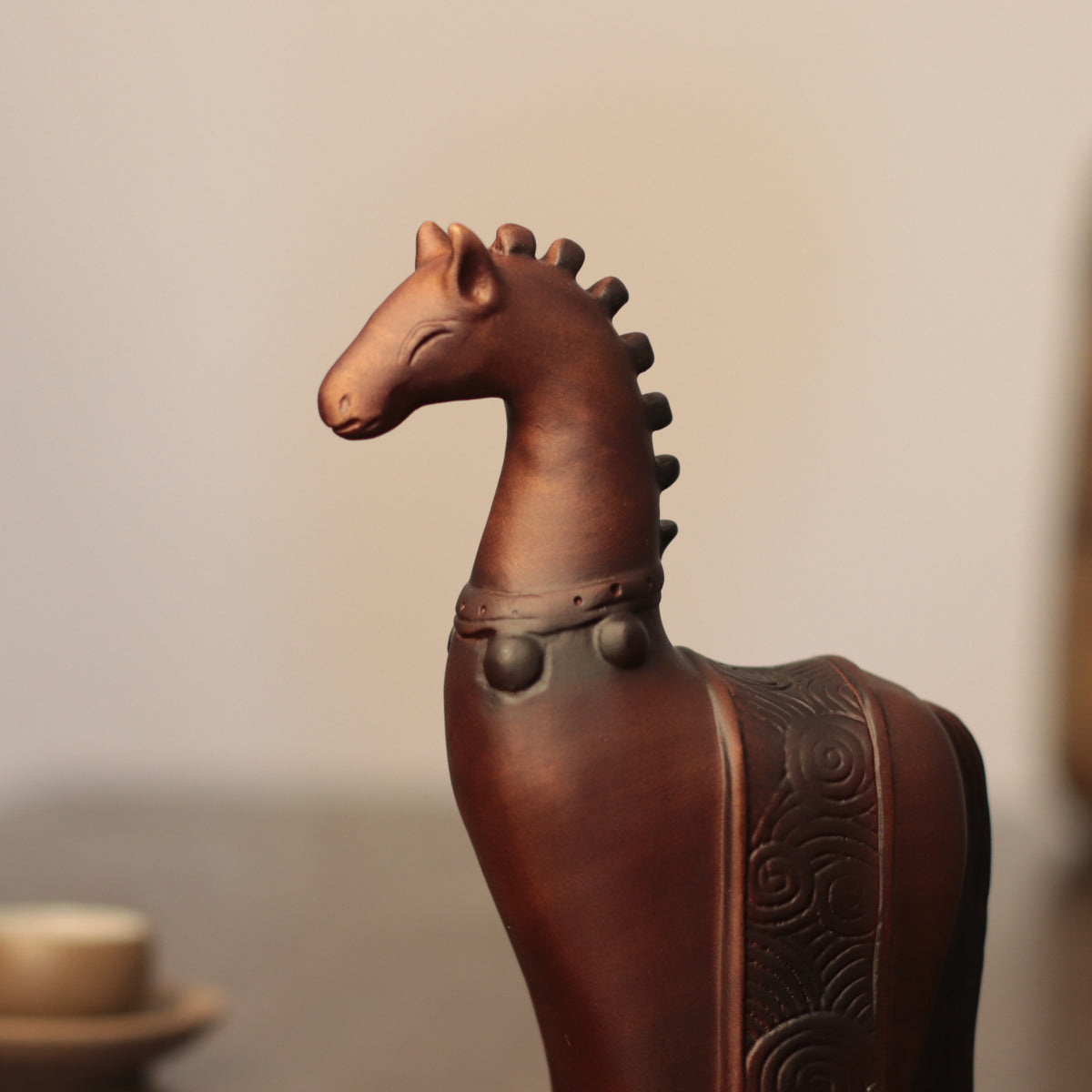 A dark brown ceramic horse figurine with carved scrollwork on its back, shown in a close-up side profile against a softly blurred neutral background.