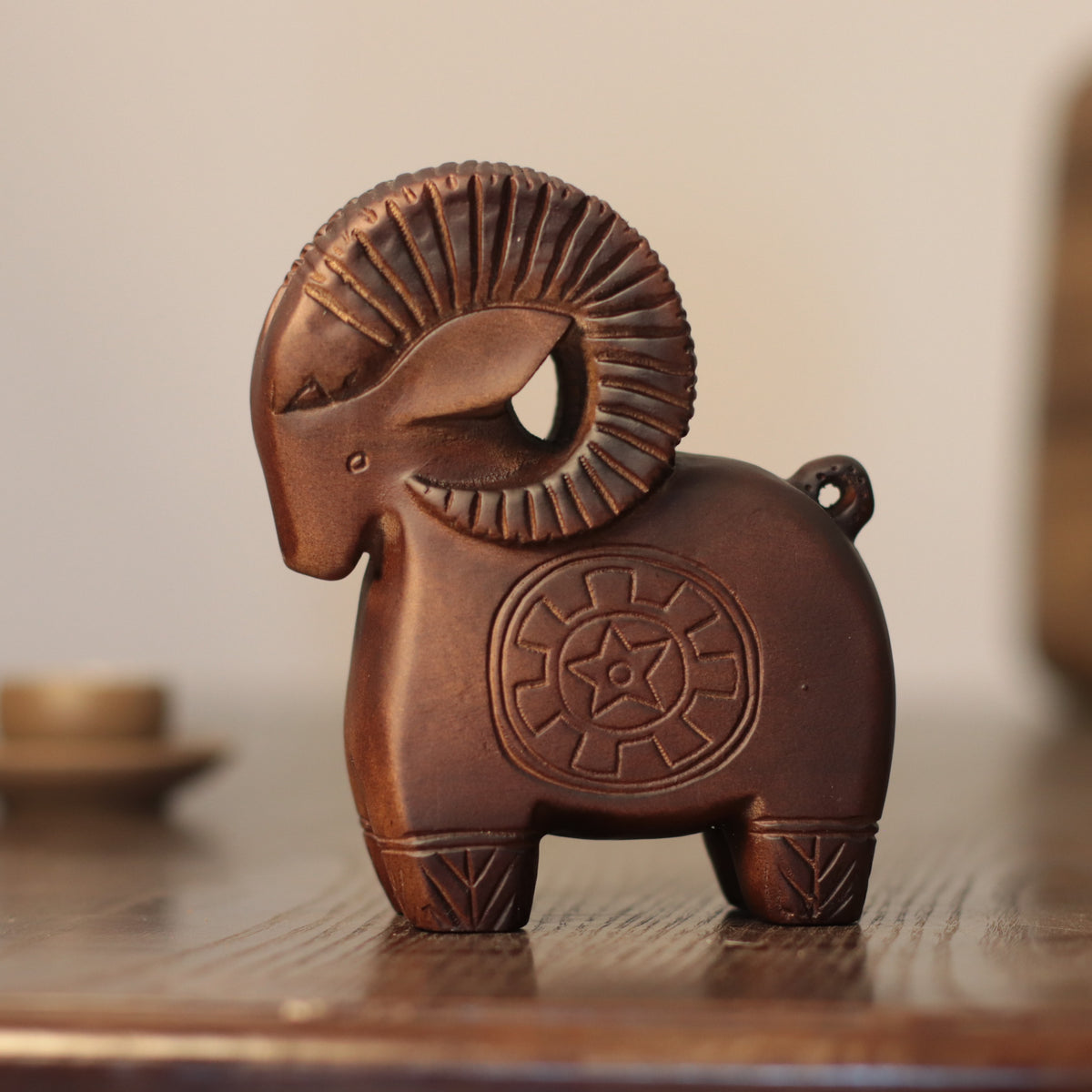 A carved wooden ram figurine with a textured, ribbed ram horn and a circular star emblem on its flank, displayed on a wooden surface against a softly blurred background.