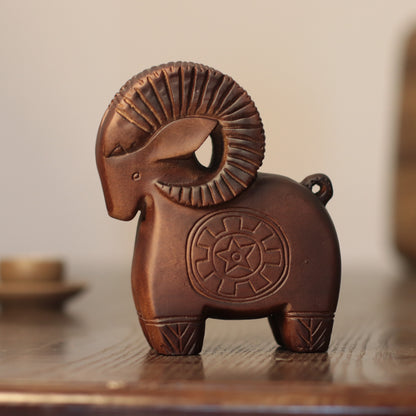 A carved wooden ram figurine with a textured, ribbed ram horn and a circular star emblem on its flank, displayed on a wooden surface against a softly blurred background.