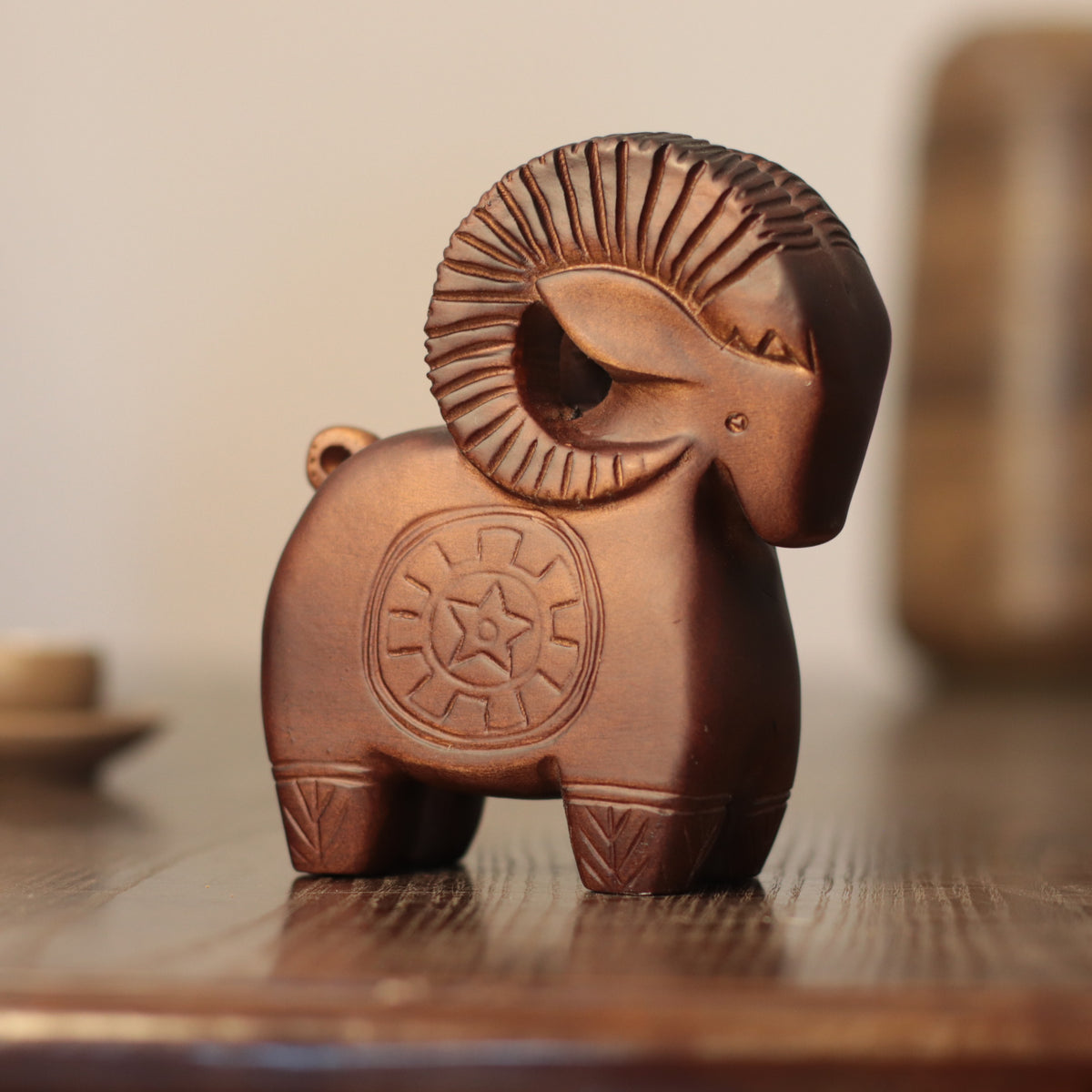A small carved ram figurine in a warm brown terracotta-like finish, standing on a wooden surface with a softly blurred indoor background.