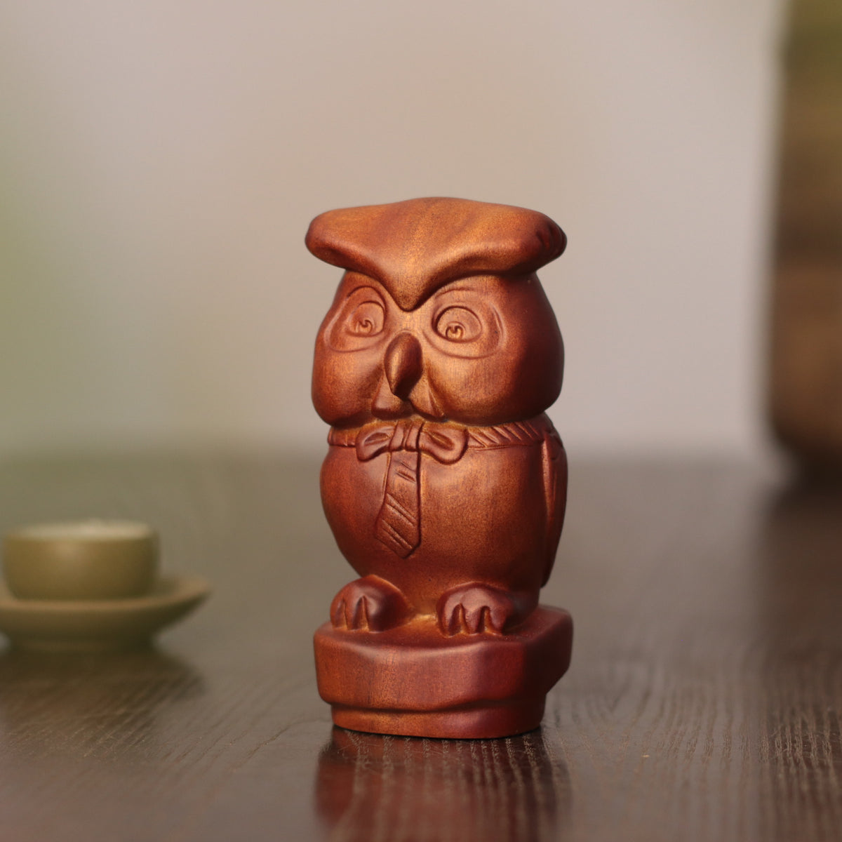 A small carved owl figurine in a warm reddish-brown wood tone, standing on a dark wooden surface with a softly blurred indoor background.