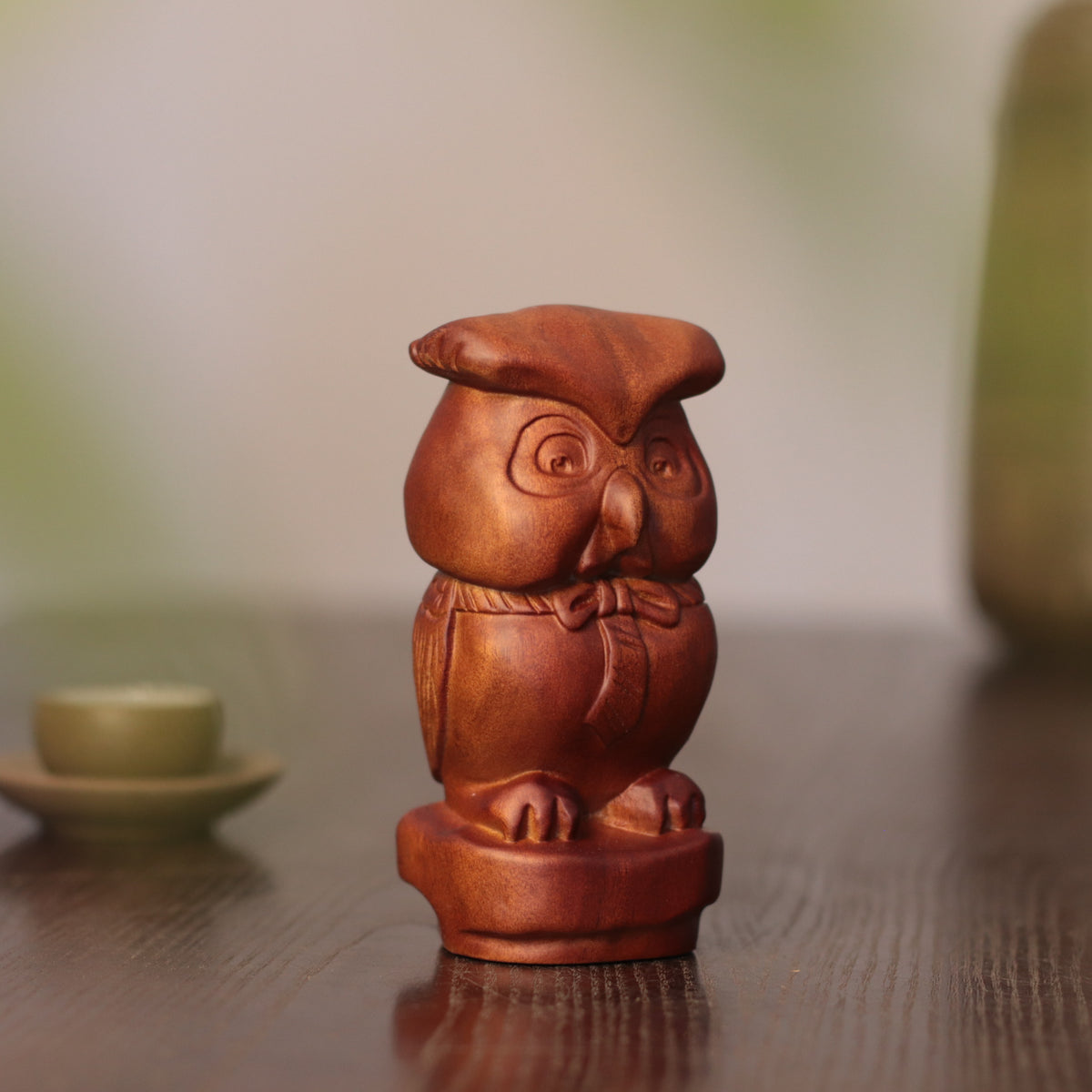 A small carved wooden owl figurine with a glossy reddish-brown finish, standing on a matching round base on a dark wooden tabletop.