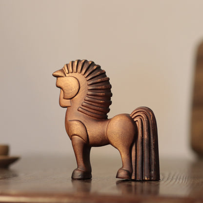 A small decorative horse figurine with a textured, metallic bronze finish, standing on a dark wooden surface against a soft, warm beige background.