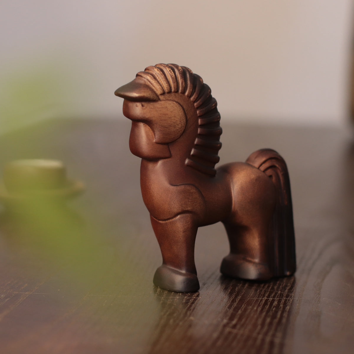 A small carved wooden horse figurine with a stylized mane, standing on a dark wooden surface against a softly blurred light background.