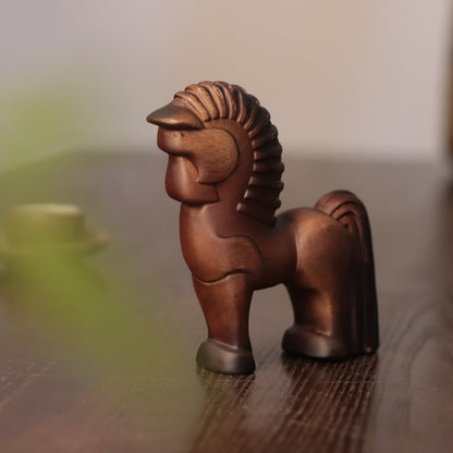 A small carved wooden horse figurine with a stylized mane, standing on a dark wooden surface against a softly blurred light background.