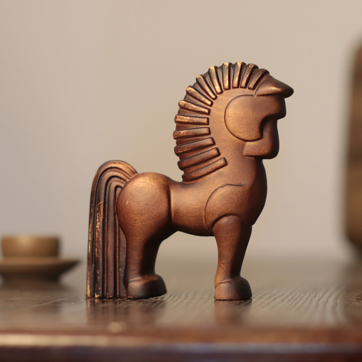 A small sculptural horse figurine in a warm metallic copper or bronze finish, standing on a wooden surface against a softly blurred neutral background.