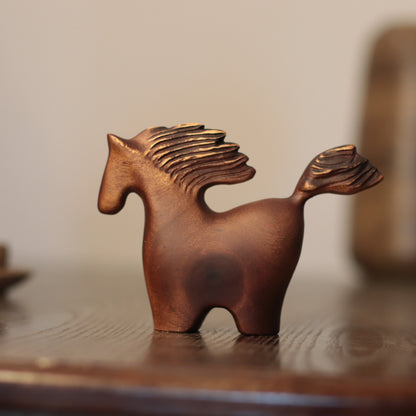 A small sculptural horse figurine in a warm copper-brown finish, standing on a dark wooden surface against a softly blurred beige background.