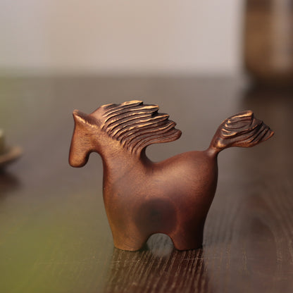 A small sculptural horse figurine in a warm metallic copper or bronze finish, standing on a dark wooden surface with a softly blurred indoor background.