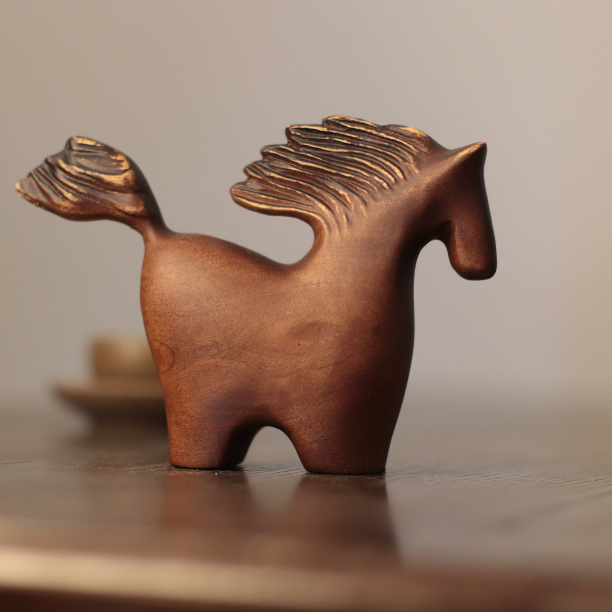 A small abstract horse figurine with a textured, metallic bronze finish, standing on a dark wooden surface against a soft, warm beige background.
