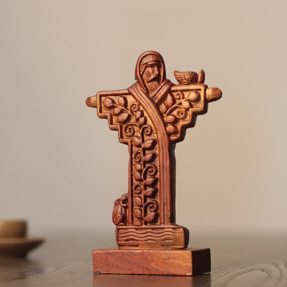 A carved wooden cross figurine of Jesus Christ with ornate scrollwork and a small bird on his shoulder, displayed on a wooden surface against a soft beige background.