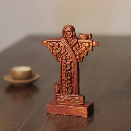 A carved wooden religious cross figurine with ornate leaf and scroll patterns, standing on a matching wooden base on a dark wooden table.