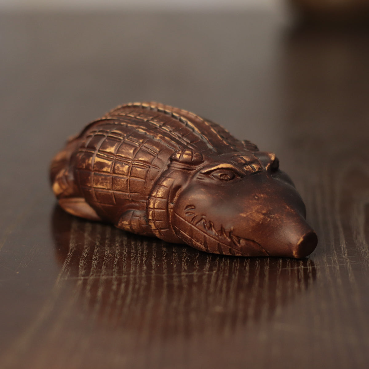 A small carved alligator or crocodile figurine with a metallic bronze finish, resting on a dark wooden surface.
