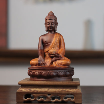 A carved wooden Buddha statue seated in a meditative pose on a decorative pedestal base, photographed indoors against a softly blurred neutral background.