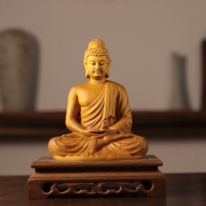 A golden Buddha statue seated in a meditative pose on a carved wooden base, photographed indoors against a softly blurred neutral background.