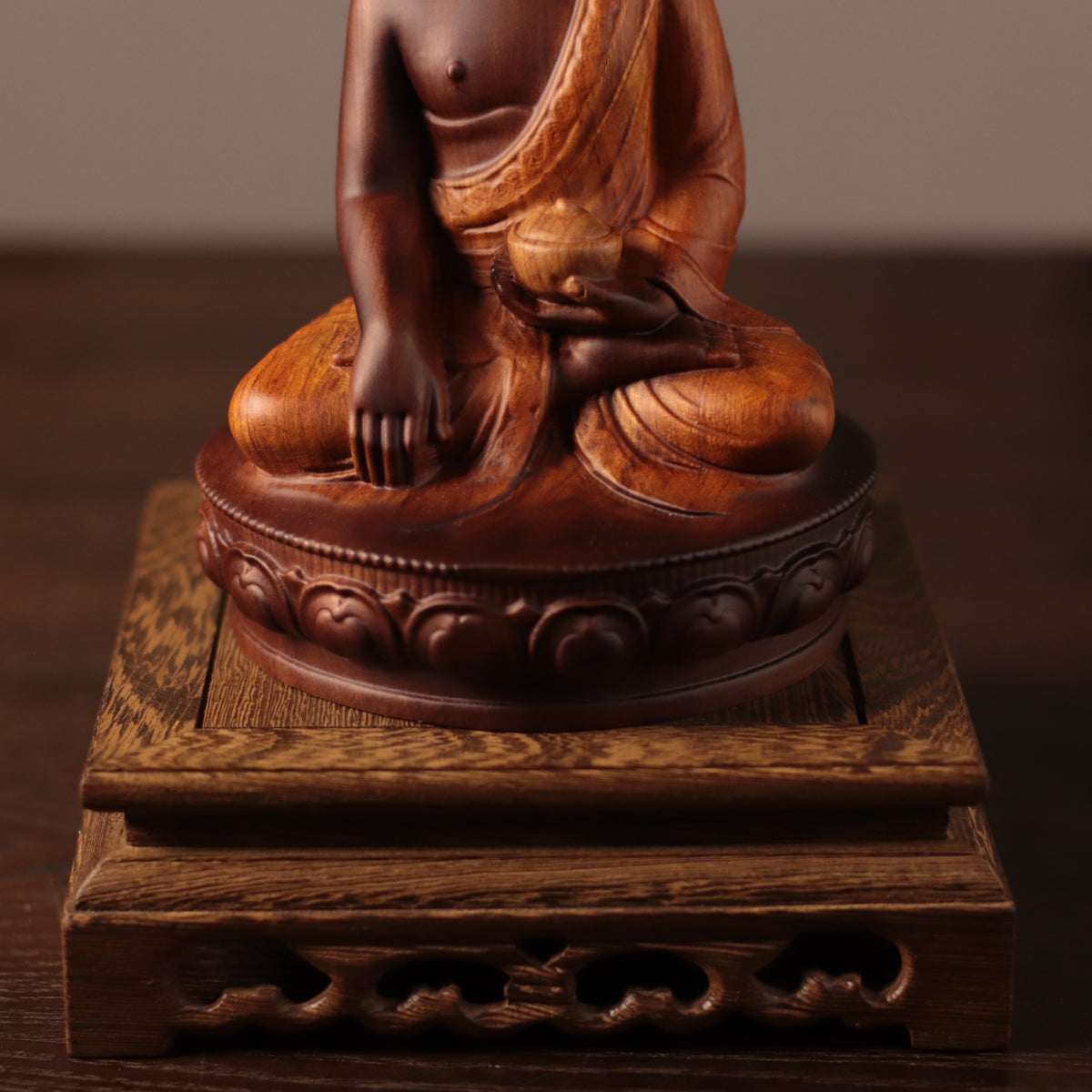 A close-up of a carved wooden Buddha statue seated on an ornate matching wooden base, shown against a dark brown background.