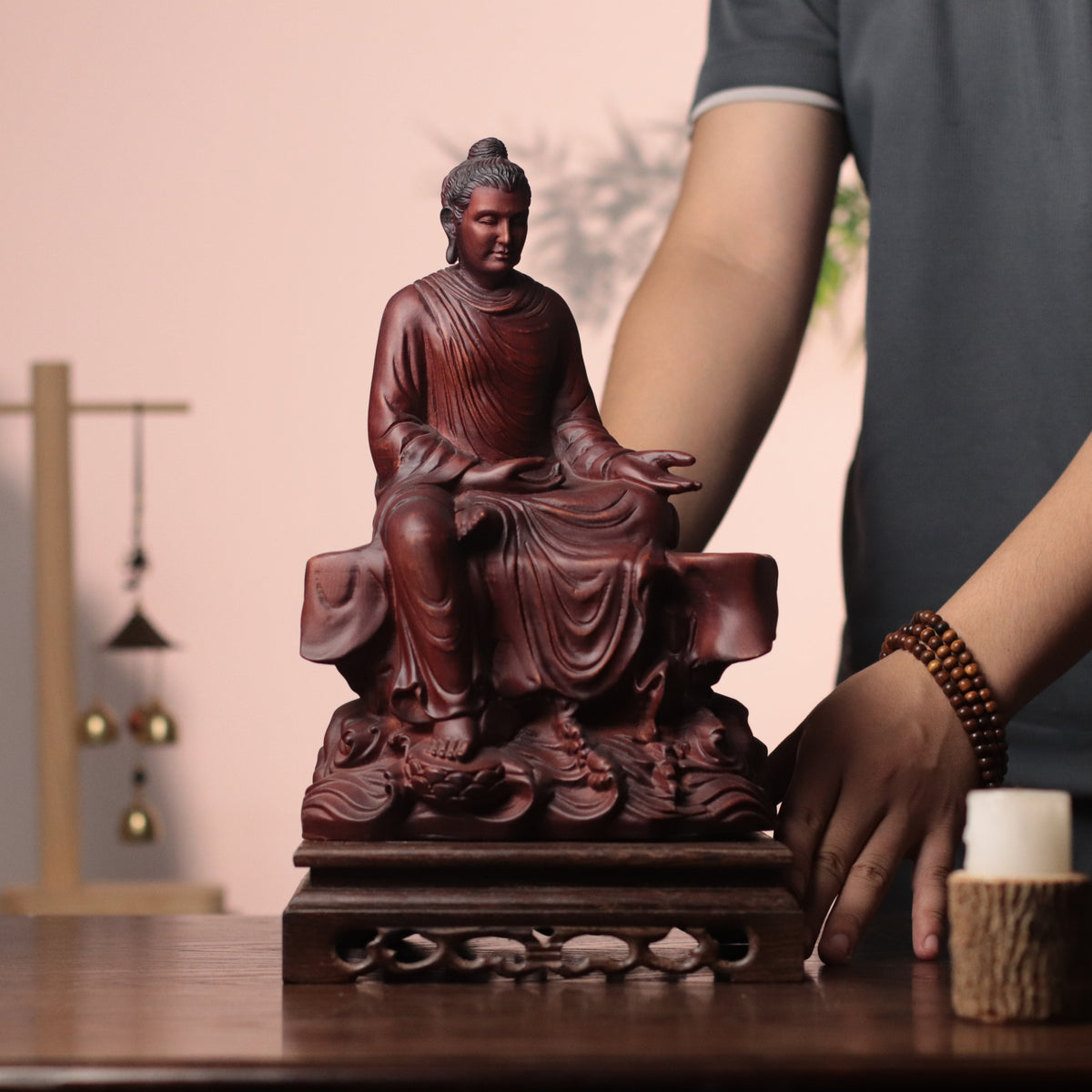 A carved reddish-brown Buddha statue seated on a pedestal, displayed on a wooden table with a person's hands and torso partially visible in a softly lit indoor setting.
