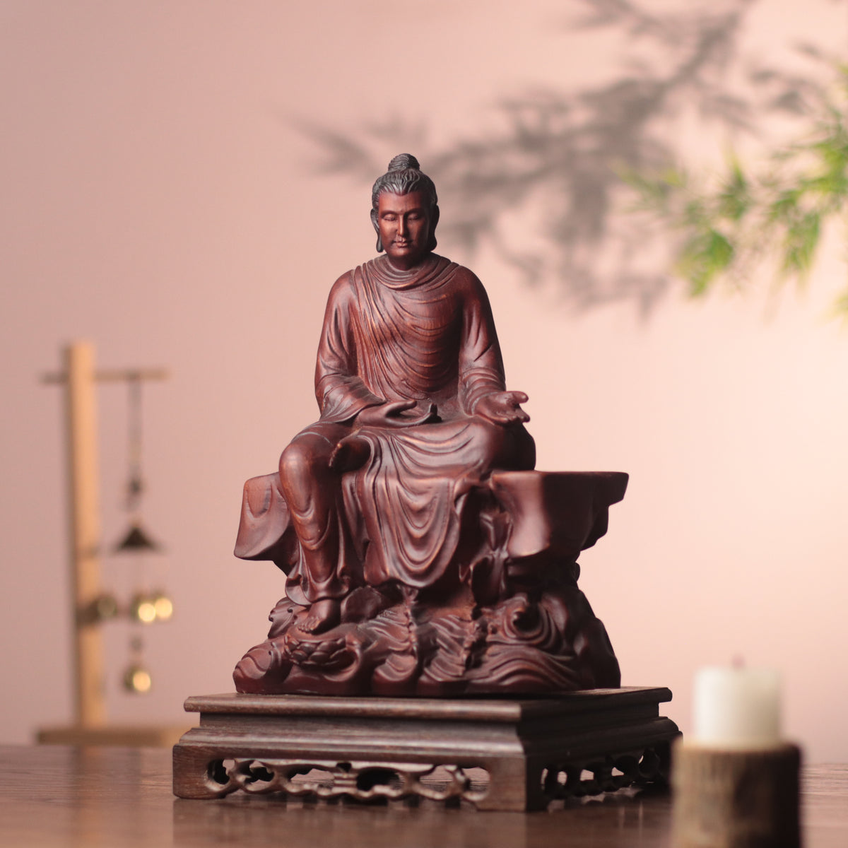 A carved Buddha statue in a reddish-brown wood finish, seated on a decorative base against a soft beige wall with a candle and hanging ornaments in the background.