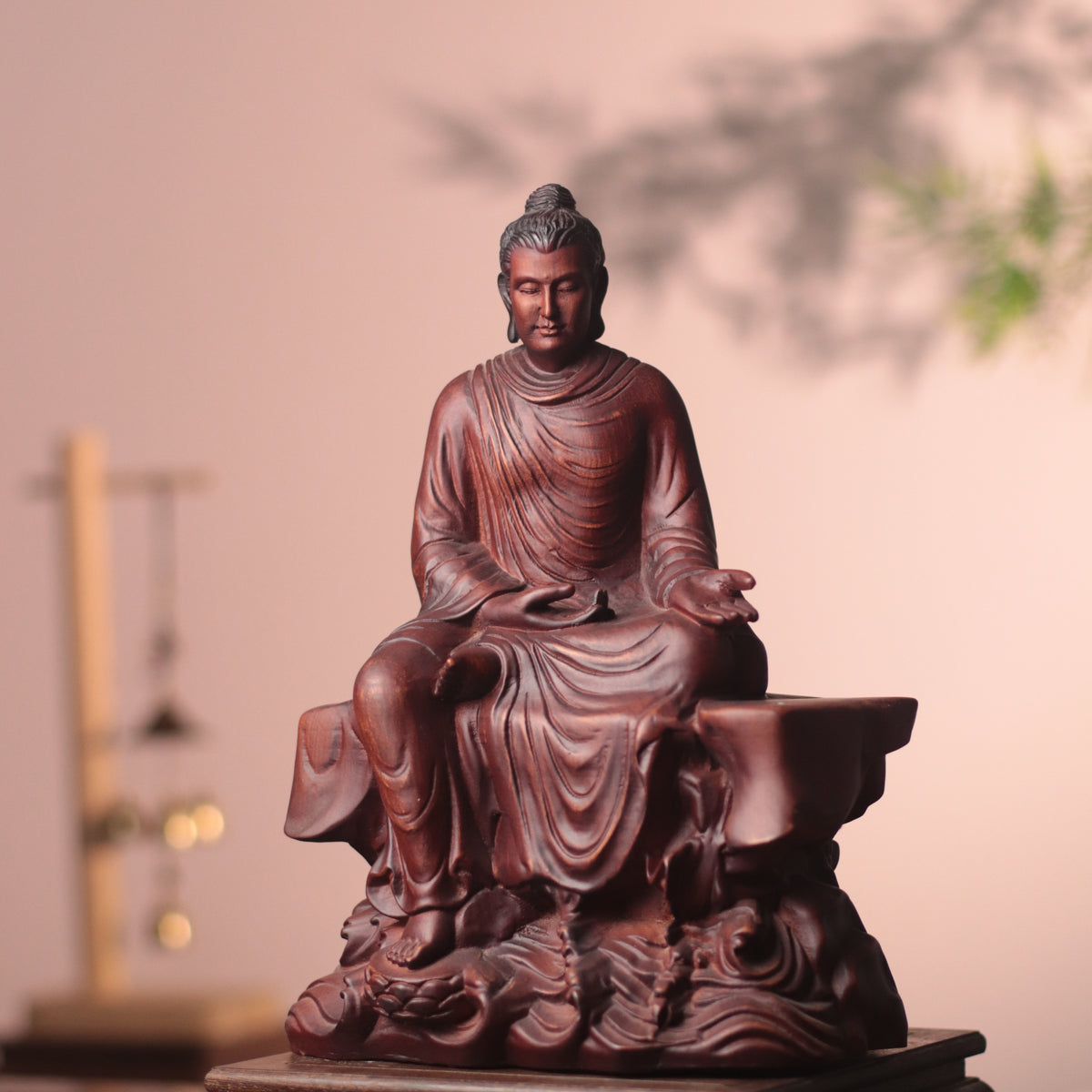 A dark reddish-brown carved Buddha statue seated on a pedestal, photographed indoors against a soft beige wall with blurred decorative elements in the background.
