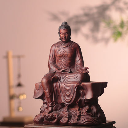 A dark reddish-brown carved Buddha statue seated on a pedestal, photographed indoors against a soft beige wall with blurred decorative elements in the background.