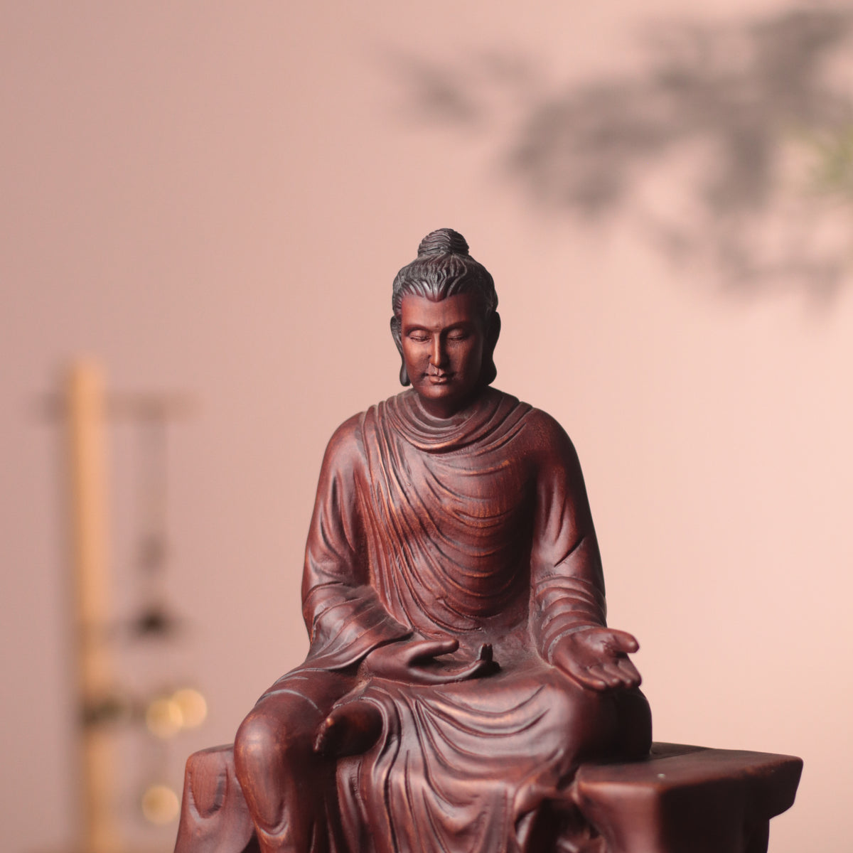 A dark reddish-brown carved Buddha statue seated cross-legged, photographed against a softly blurred warm beige background with a faint tree shadow.