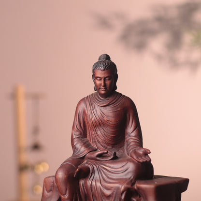 A dark reddish-brown carved Buddha statue seated cross-legged, photographed against a softly blurred warm beige background with a faint tree shadow.